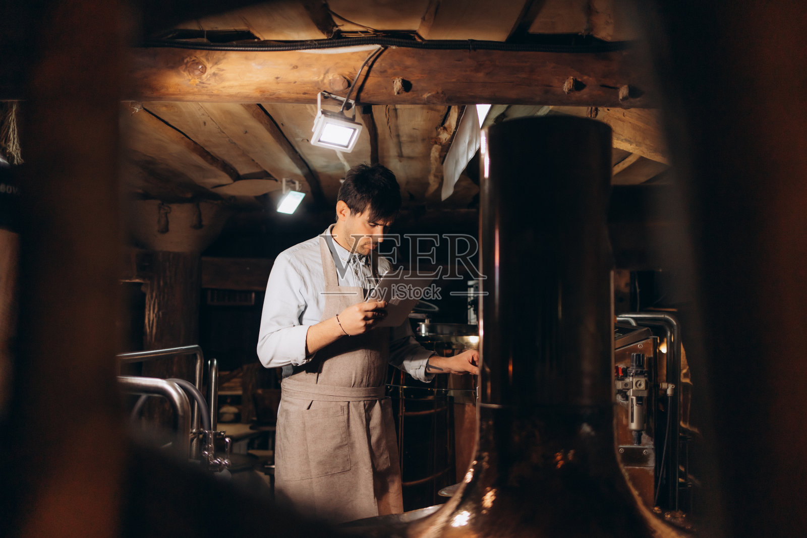 啤酒质量控制。戴着围裙和防护面具的男子拿着数码平板电脑看着啤酒厂的啤酒杯，自由的空间照片摄影图片