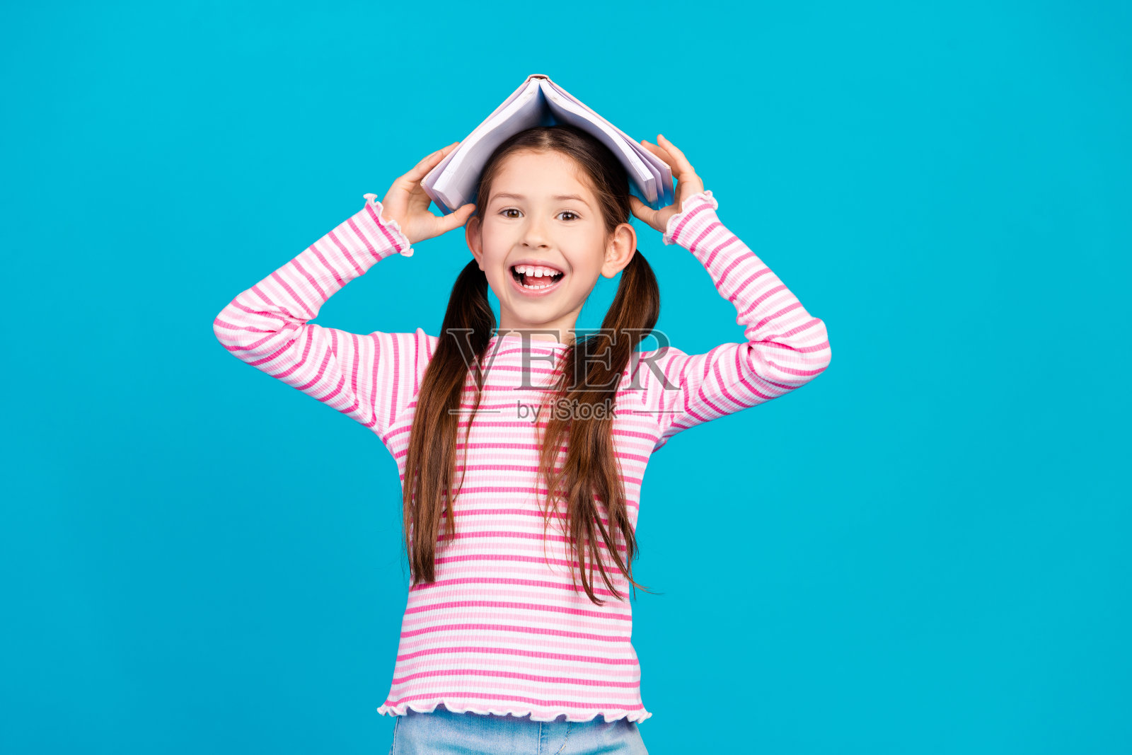 照片的有趣的积极的小女孩与长马尾辫穿着条纹衬衫拿着书的头孤立的蓝色背景照片摄影图片