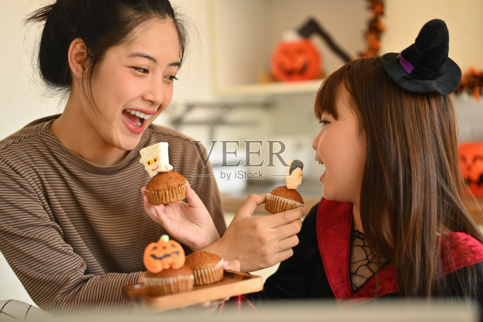 可爱的小女孩打扮成女巫和妈妈在厨房吃万圣节纸杯蛋糕。人，节日和节日的概念照片摄影图片