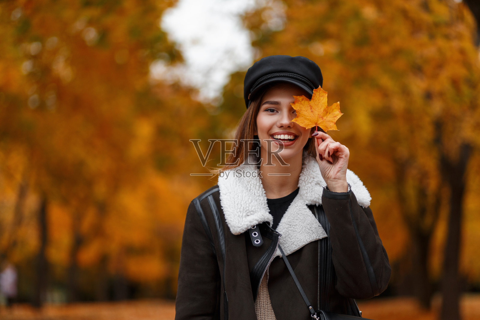 一个非常乐观的年轻女子，戴着一顶时髦的黑帽子，穿着一件时髦的棕色夹克，在公园里拿着一片黄橙色的枫叶，微笑着。有趣的女孩在秋天的森林里享受周末。照片摄影图片