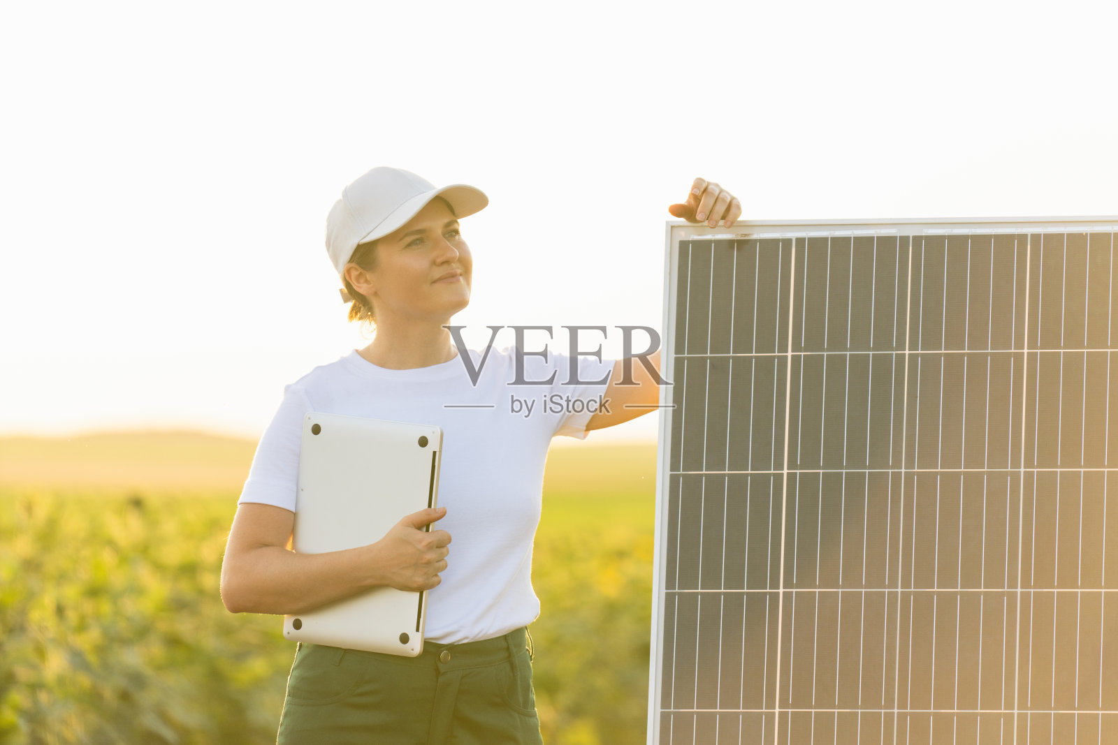 戴着白色帽子、穿着t恤、拿着笔记本电脑的农妇站在太阳能电池板旁边照片摄影图片