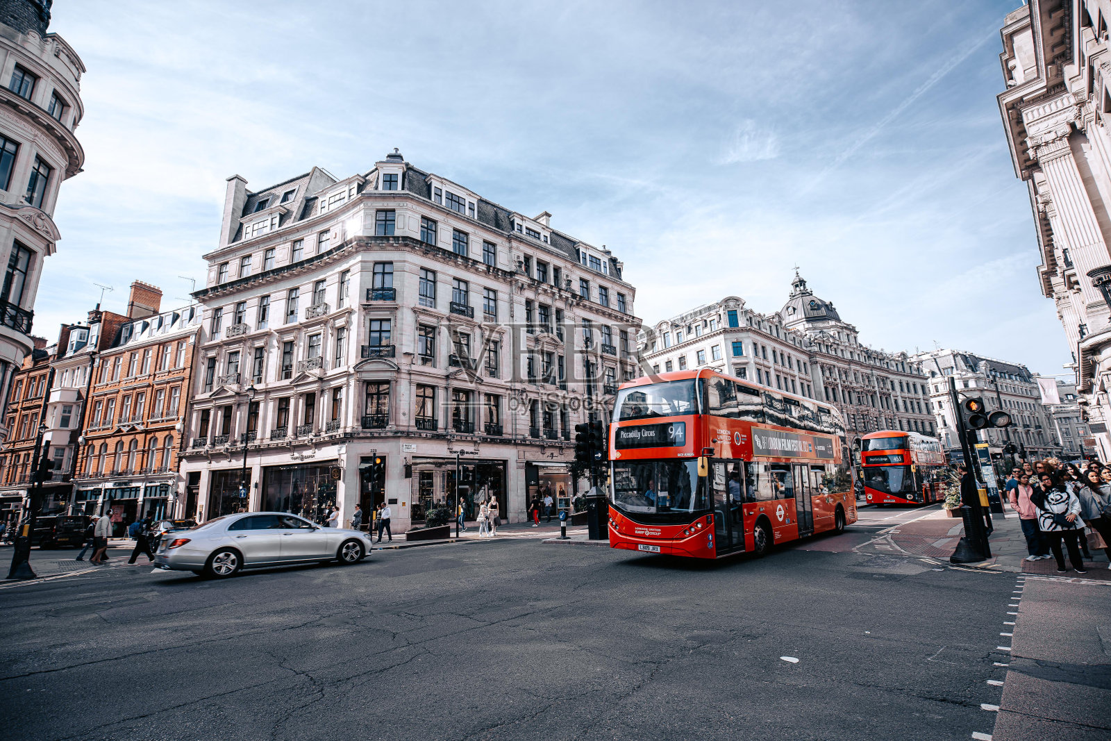 一辆红色的双层巴士沿着伦敦的街道行驶,经过行人的人行横道,一辆银色的汽车正在行驶。公共汽车上有个牌子写着“皮卡迪利广场”。照片摄影图片