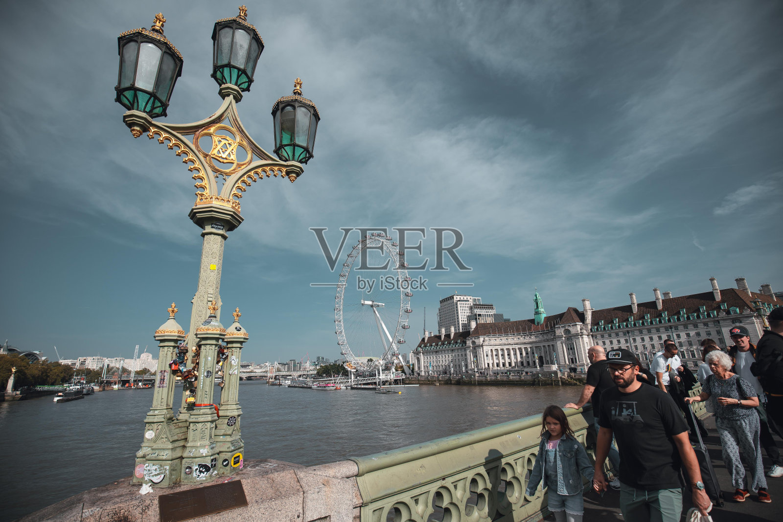 从泰晤士河上的一座桥上俯瞰伦敦眼,行人走过装饰着锁的灯柱。照片摄影图片