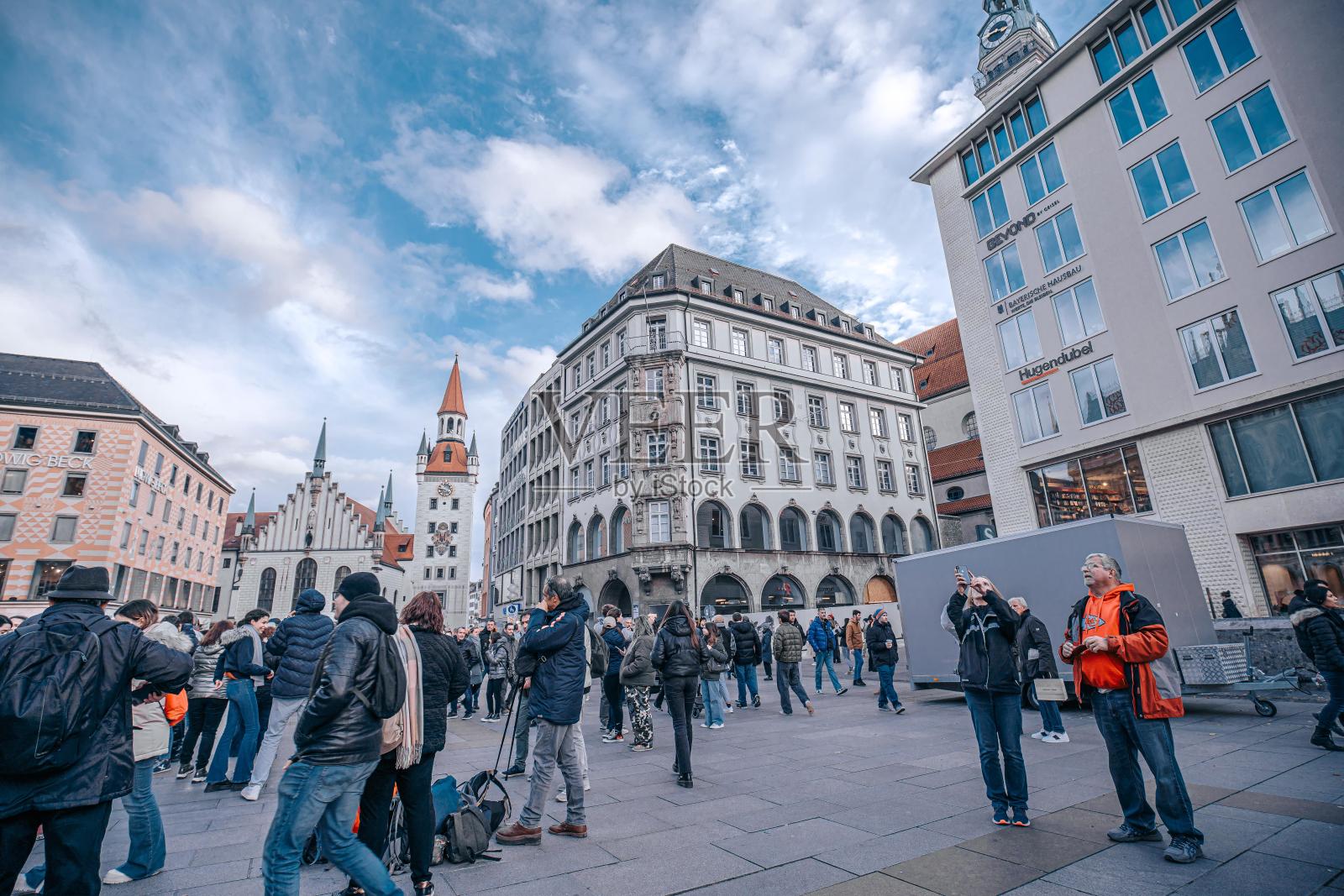在德国慕尼黑，人们聚集在一座建筑物前，背景是一座高耸的钟楼。照片摄影图片