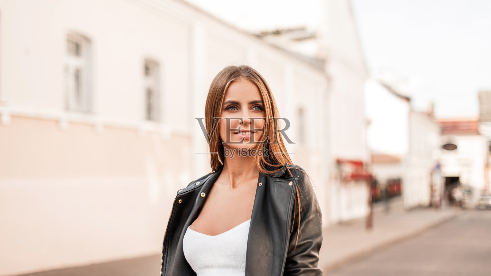 一个美丽的年轻女子的肖像，带着可爱的微笑，自然的妆容，棕色的头发，时尚的黑色皮夹克和白色优雅的上衣。迷人的快乐女孩走在城市的街道上照片摄影图片