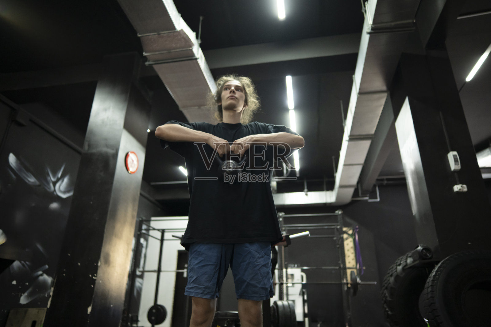 一个留着长发的年轻人在一个黑色墙壁和管道的现代健身房锻炼时举着壶铃照片摄影图片