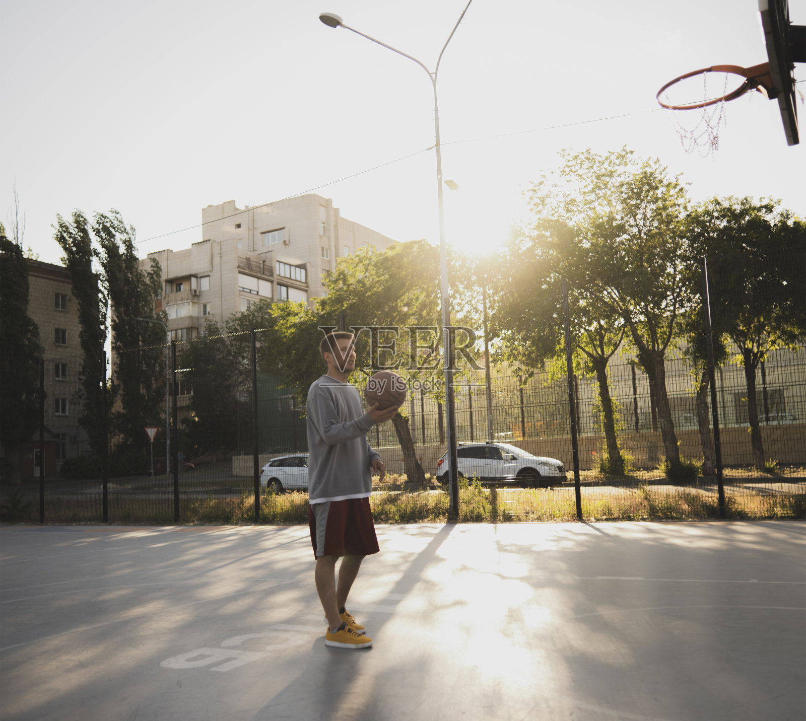 一个年轻人站在篮球场上，手里拿着一个篮球。太阳在他头顶上明亮地照耀着，在球场上投下了温暖的光芒，在背景的树上形成了长长的阴影照片摄影图片