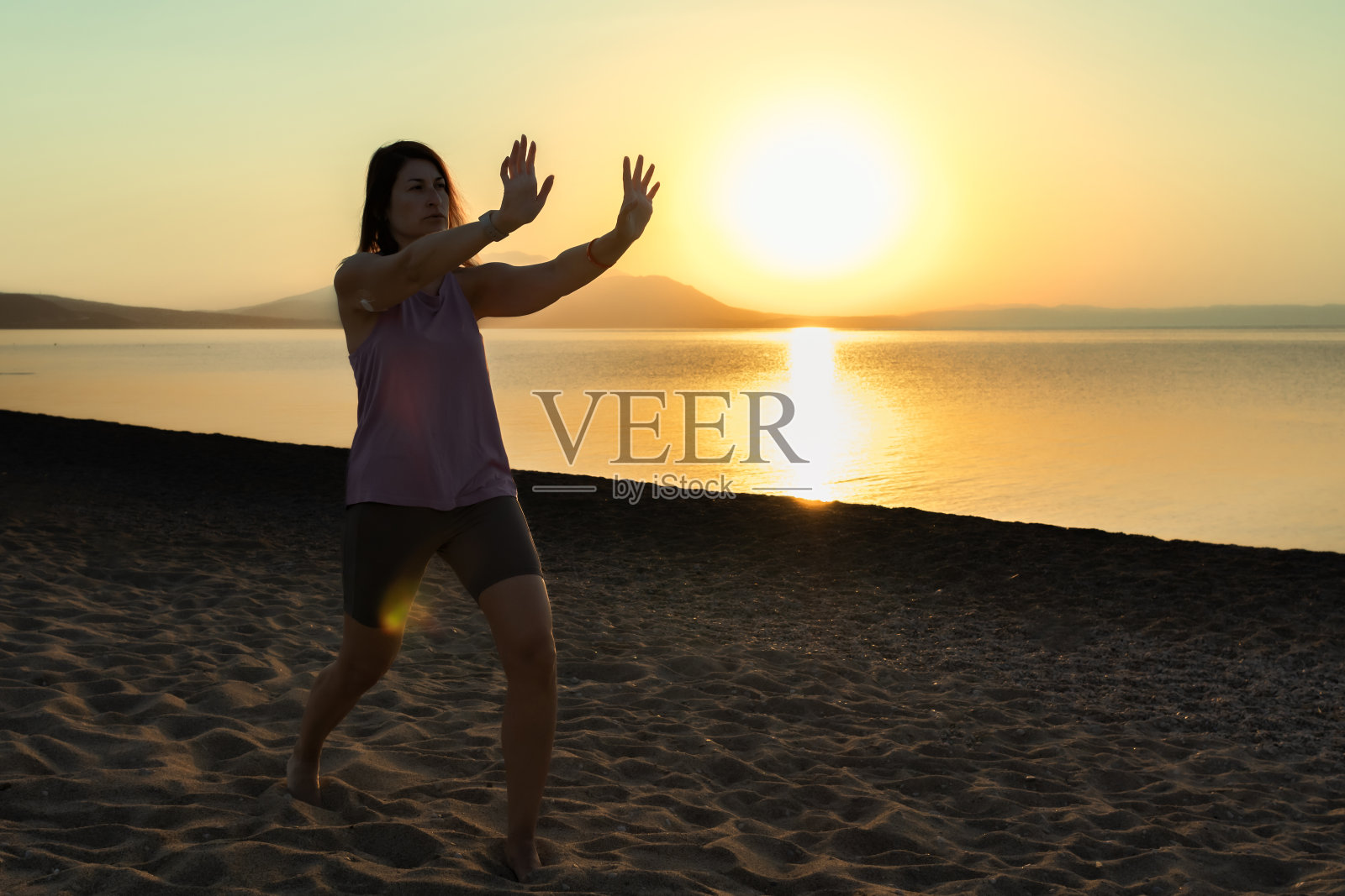 中年糖尿病妇女在日落或日出时在海边的沙滩上练习太极。轮廓。健康、积极的生活方式。运动与糖尿病。一位女士在上臂背部佩戴了控制血糖的小传感器。照片摄影图片