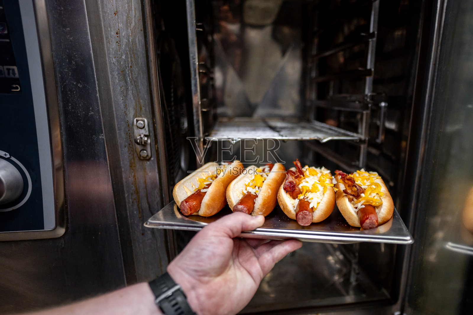 手拿着一个装有四个热狗的托盘，站在一个敞开的工业烤箱前照片摄影图片