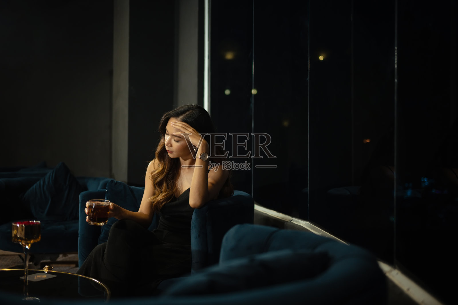 沉思的年轻成年女子独自坐在灯光昏暗的休息室里，手里拿着一杯饮料，陷入了沉思照片摄影图片