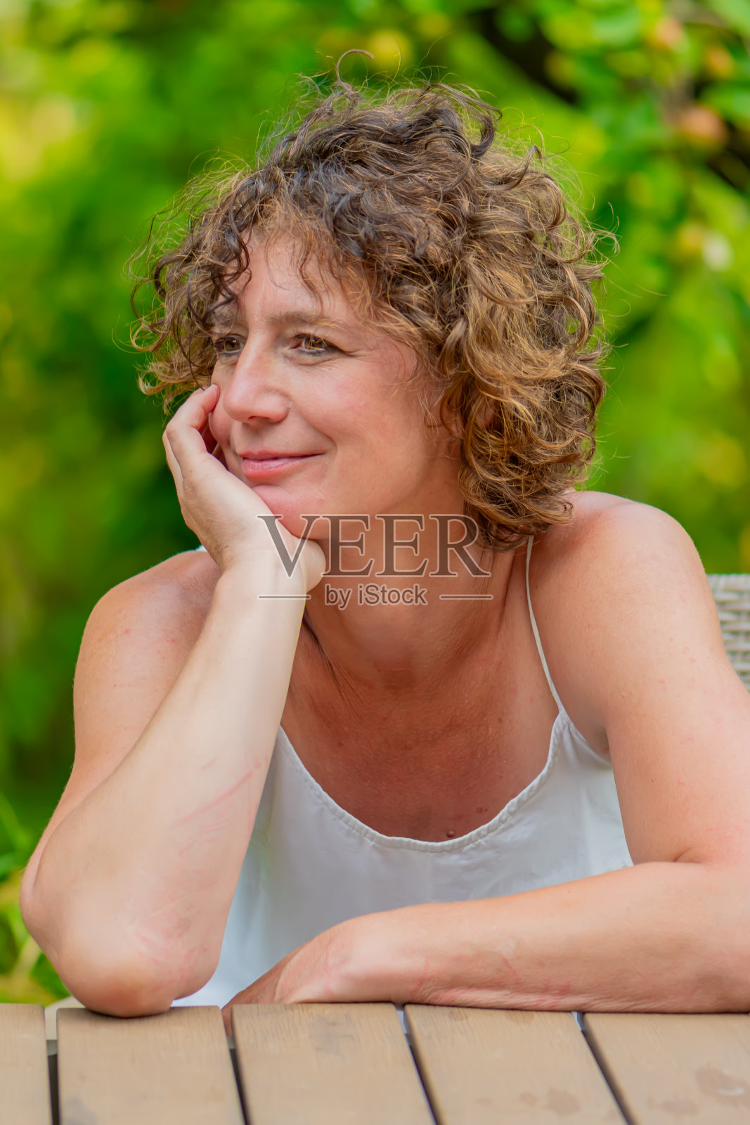 一个棕色卷发的女人坐在户外的桌子旁，用手托着下巴，沉思地凝视着远方。照片摄影图片