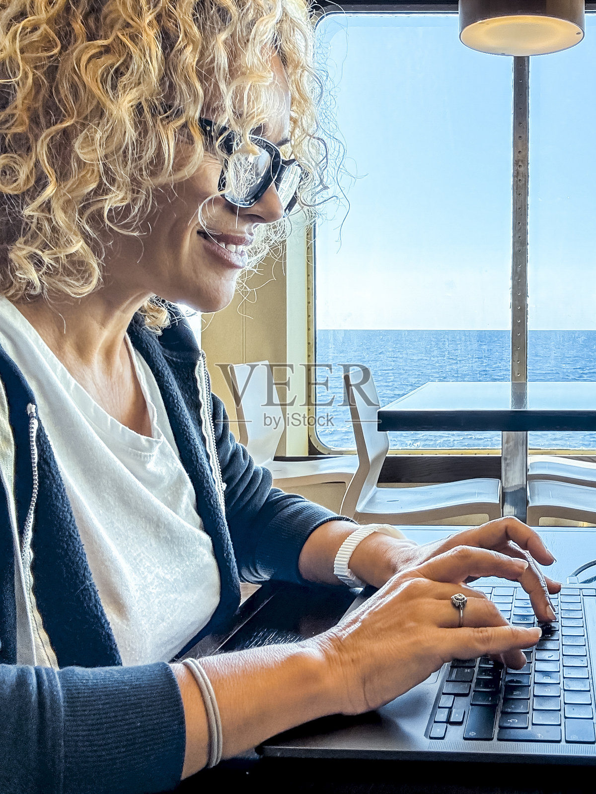 乘船旅行时坐在餐桌旁的金发美女经理。卷发戴黑眼镜的女人在旅行时使用笔记本电脑。数字游牧民的概念。照片摄影图片