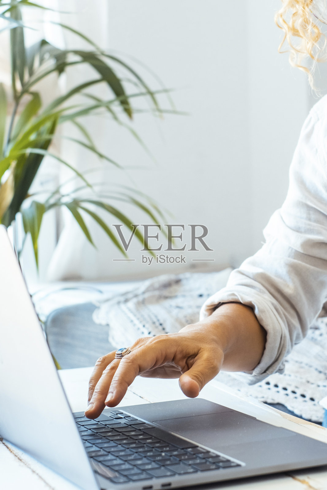 金发卷发的女人坐在家里的沙发上用笔记本电脑。迷人的中年女士一边工作，一边拿着一卷未完成的项目，并在笔记本电脑上检查她的工作。照片摄影图片