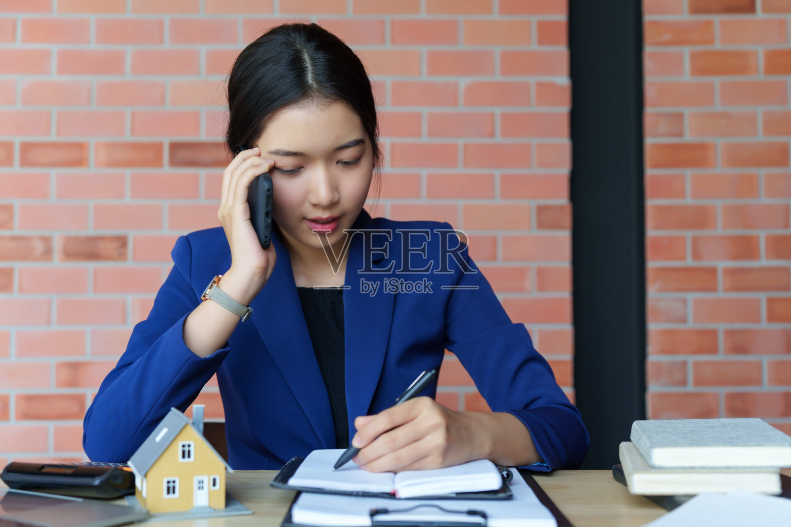 一位身穿蓝色西装的女商人坐在一张以砖墙为背景的办公桌前。她一边打电话，一边在笔记本上写东西照片摄影图片