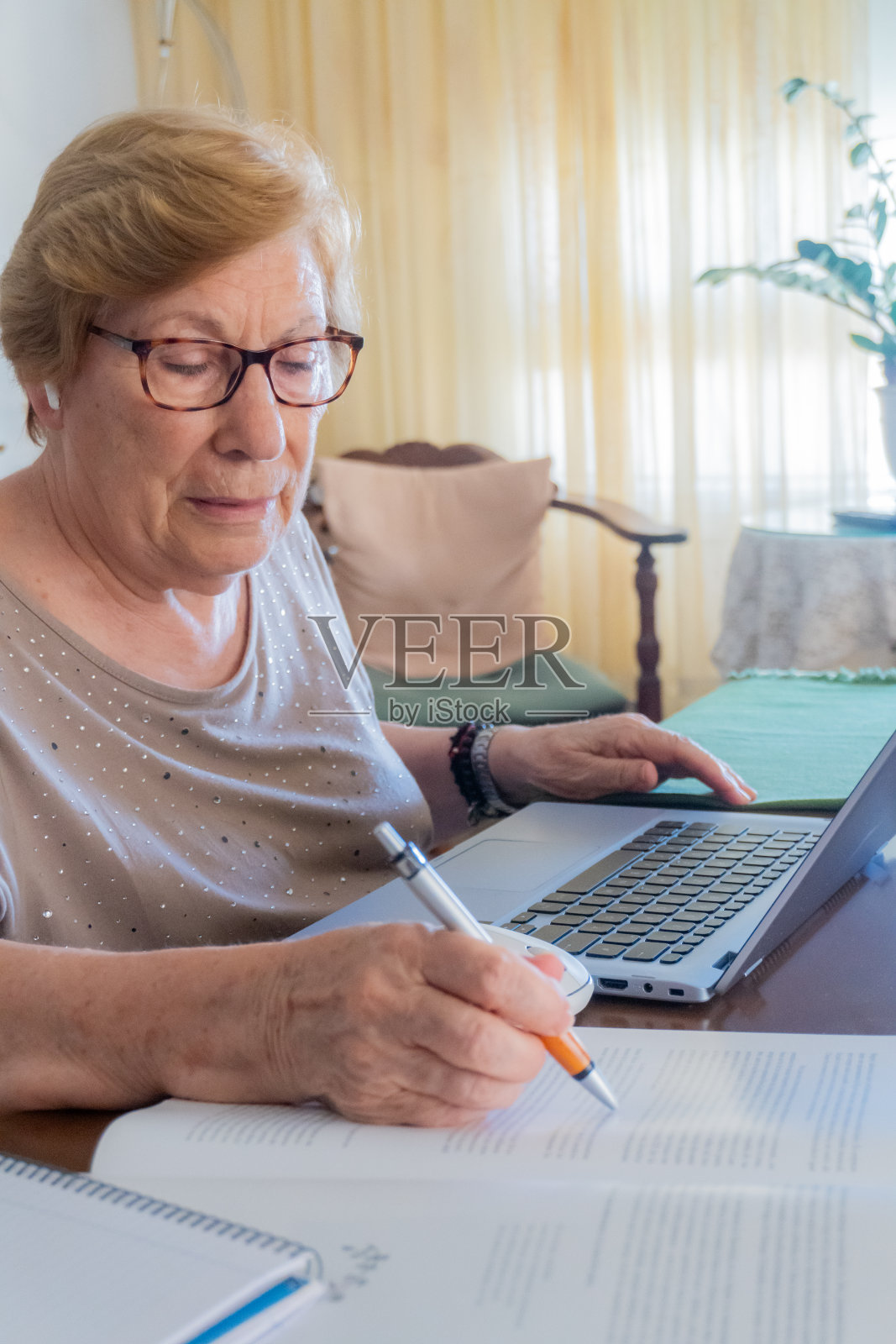 一位老妇人在家里用笔记本电脑和一本书学习。照片摄影图片