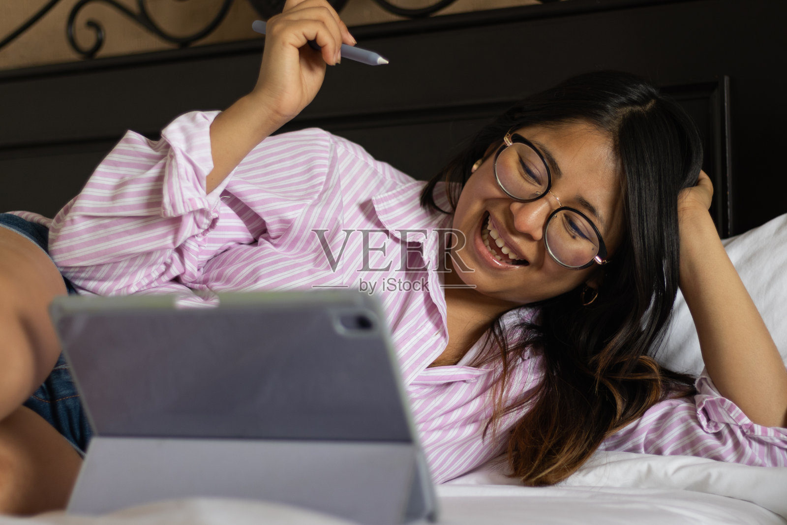 一名年轻女子微笑着躺在酒店的床上，手里拿着电子笔，在卧室里看着她的平板电脑上网照片摄影图片
