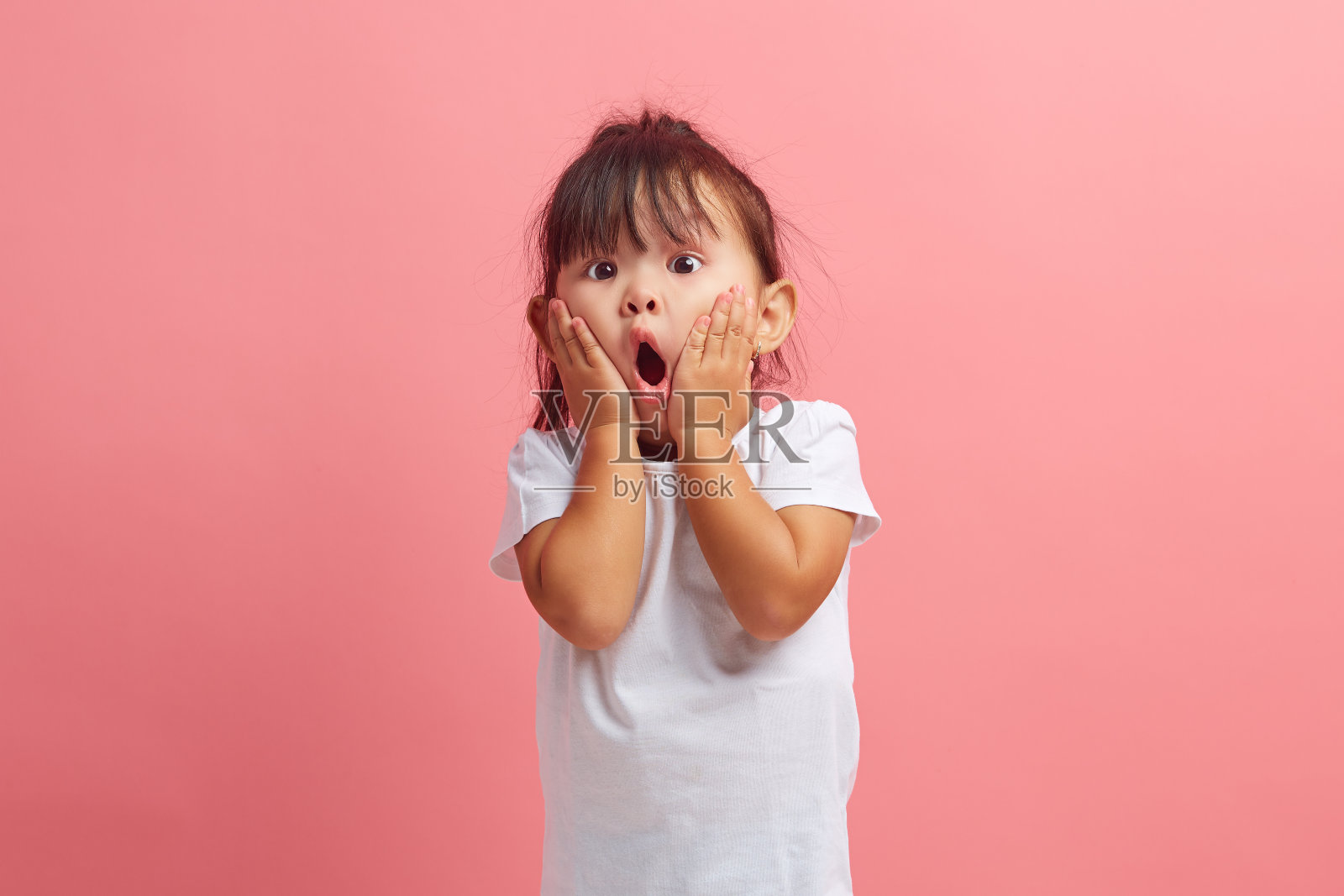 三岁的小女孩惊讶地张开嘴双手放在脸颊上震惊地盯着镜头孤立在粉红色