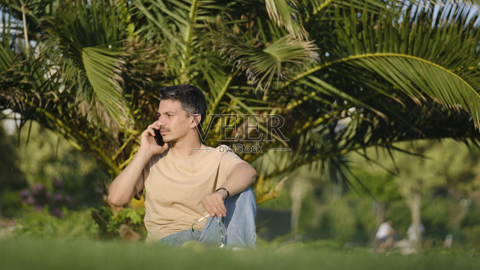 一个英俊、年轻、黑发的男人躺在一个树木繁茂的公园的草地上，一边玩一边打电话照片摄影图片