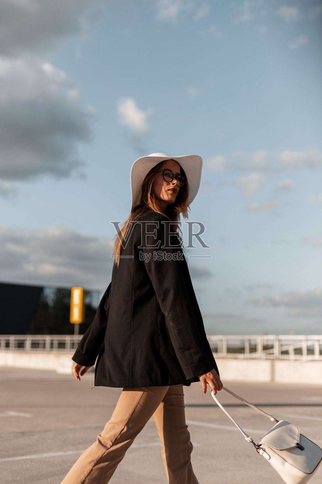 时尚的年轻女子，穿着时尚的黑色运动上衣，戴着时尚的草帽，穿着优雅的裤子，挎着包，在阳光明媚的日子里走在城市的停车场上。新的时尚休闲服装的妇女。照片摄影图片