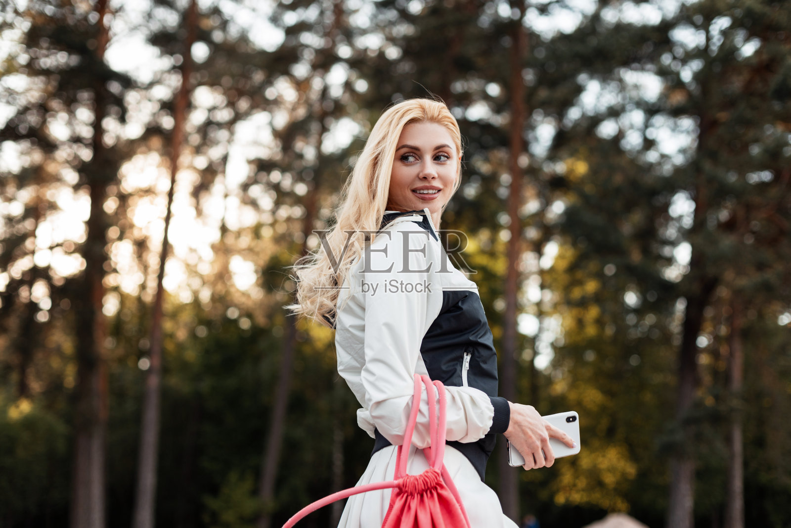 年轻的金发女郎穿着运动时尚的黑白夹克，背着粉红色时髦的背包，微笑着在夕阳下的森林里摆姿势。积极的女孩时尚模特带着微笑，穿着时髦的运动服走在公园里。照片摄影图片