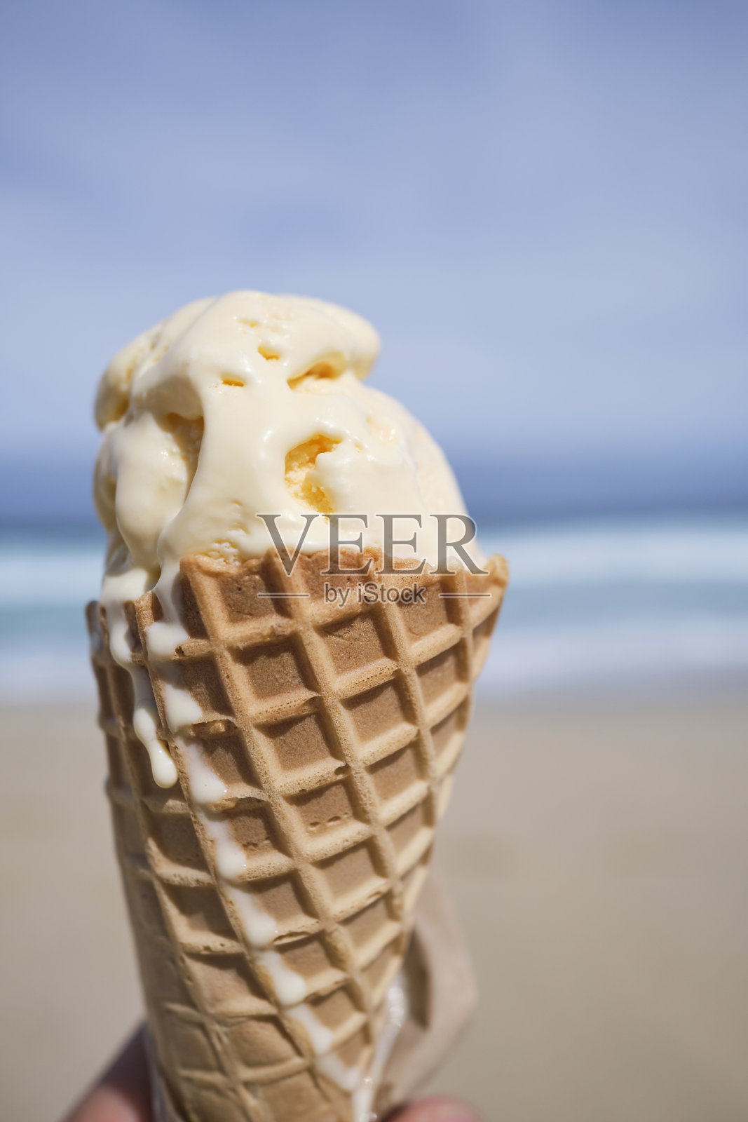 6月的一个阳光明媚的日子，一只手在康沃尔郡圣艾夫斯的波思格维登海滩的海岸线上拿着香草冰淇淋甜筒。照片摄影图片