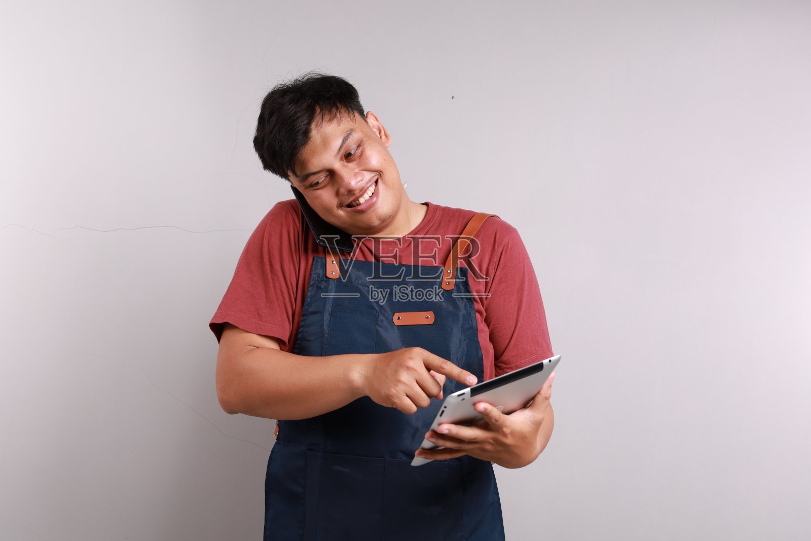 年轻的亚洲男子咖啡师调酒师酒保员工穿着蓝色围裙在咖啡店工作使用平板电脑电脑用手机说话孤立在白底小型企业创业照片摄影图片