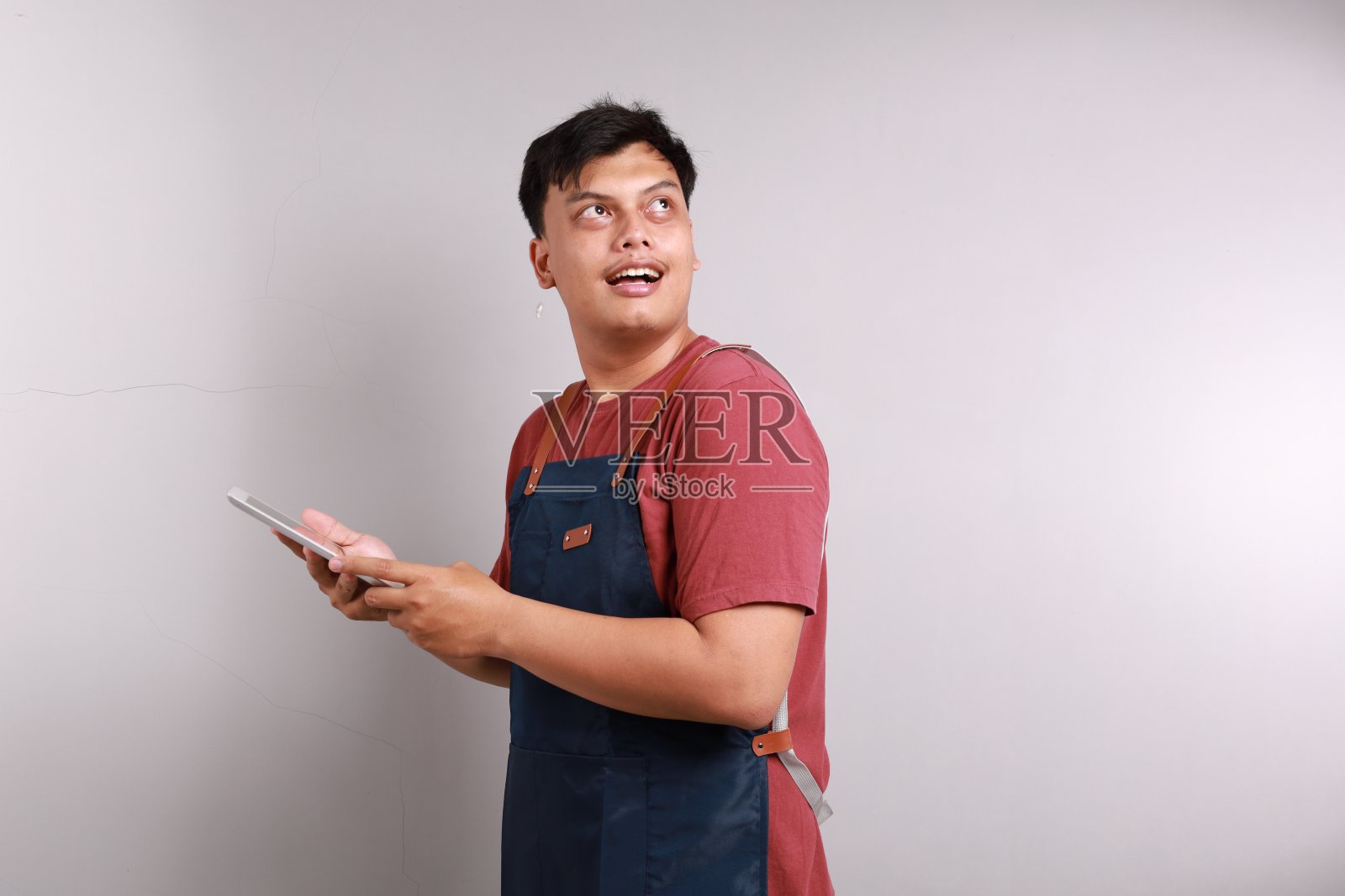 微笑的年轻亚洲人，咖啡师，酒吧服务员，穿着蓝色围裙，在咖啡店工作，拿着数码平板电脑，看着白色背景上孤立的副本空间。小型企业创业概念照片摄影图片