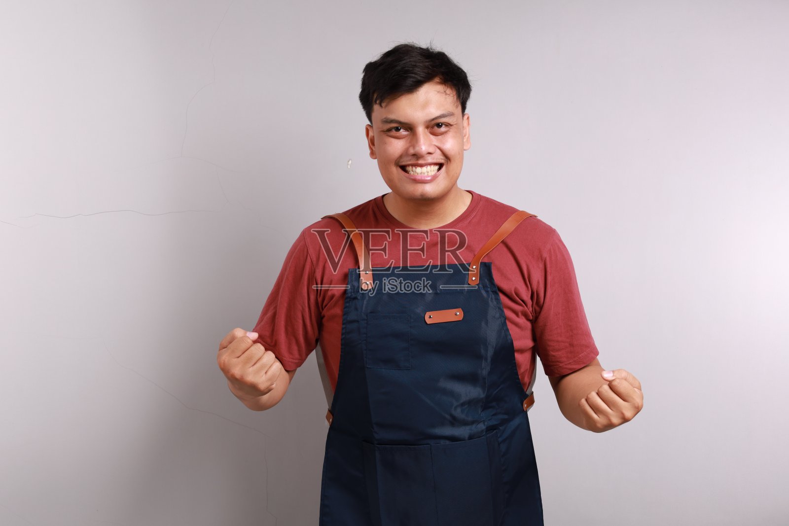 穿着蓝围裙的咖啡店老板兴奋地庆祝，挥舞拳头，欢呼雀跃，实现了目标，站在白色的背景下。照片摄影图片