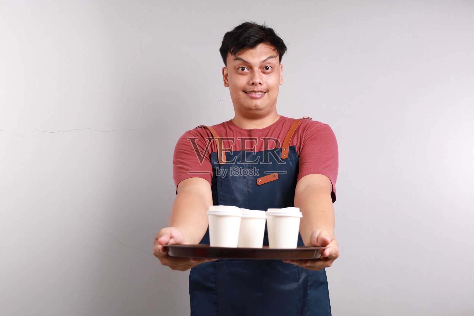 穿着蓝围裙的友好的亚洲咖啡师正在点外卖，端着咖啡，面带微笑，站在白色的背景上照片摄影图片