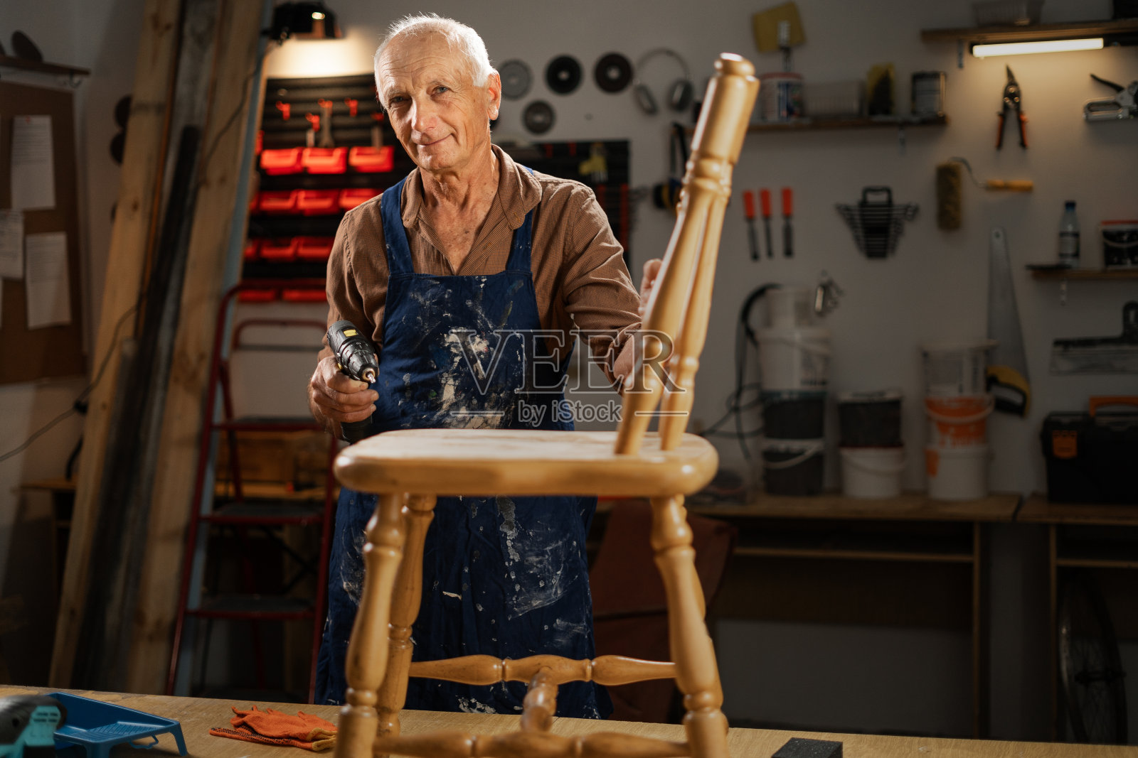 一位老人，准备在他的木工店里重新粉刷一把木椅。拿着电钻，系着染了漆的蓝色围裙，准备自己动手。照片摄影图片