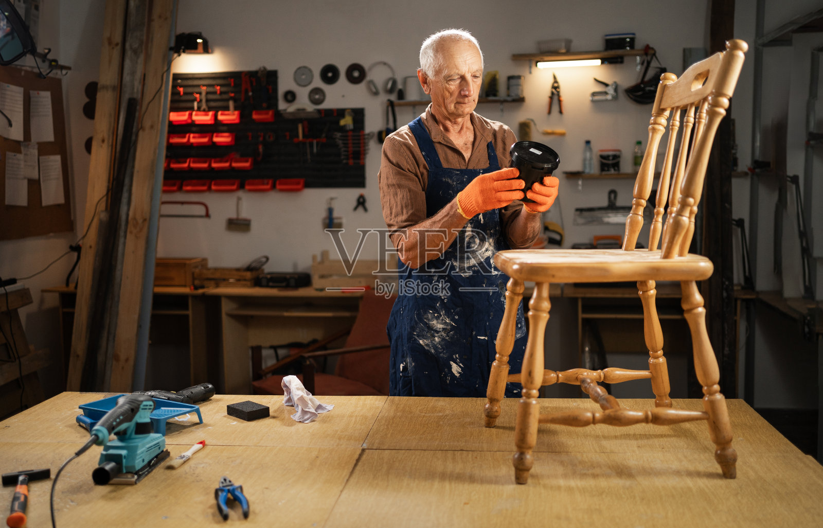 一位穿着油漆围裙的快乐老人正在看罐子上的油漆成分。站在他的工作室里。旧家具修复，概念修复和旧椅子的第二次生命照片摄影图片