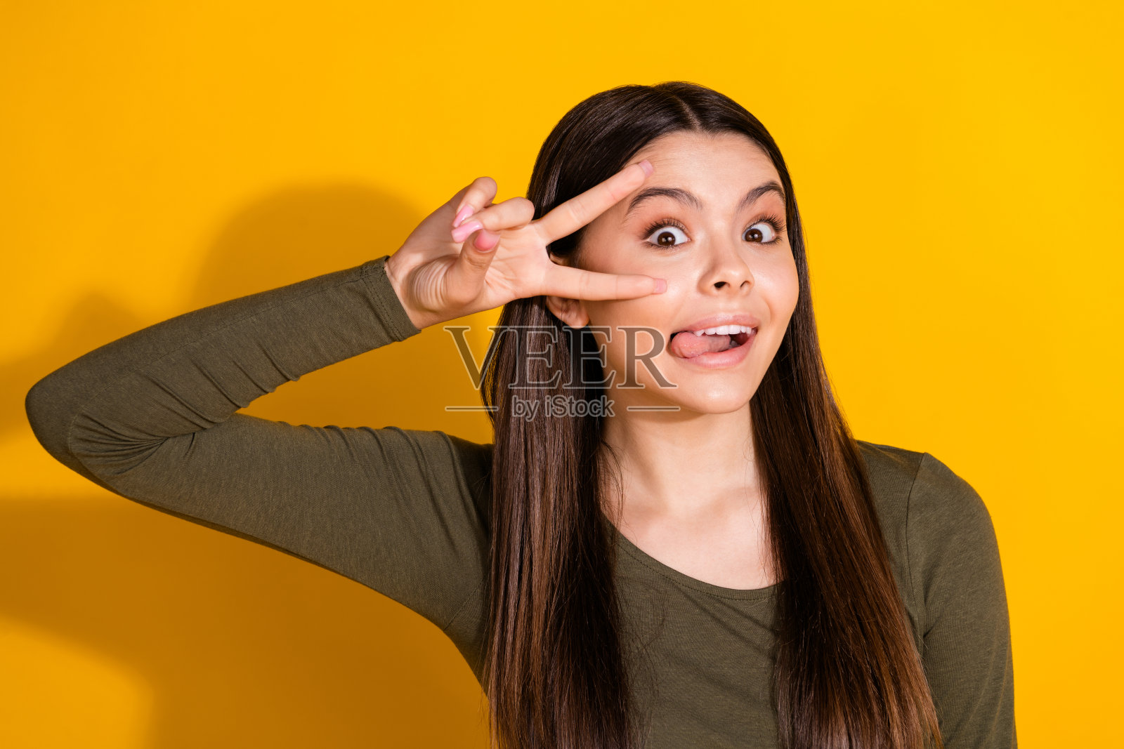 照片中，漂亮的少女露出v字舌头，穿着黄色背景的时髦卡其布服装照片摄影图片