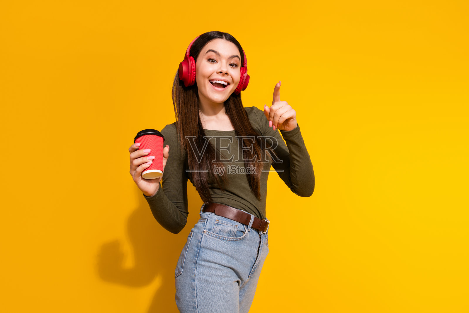 漂亮少女的照片肖像，耳机，咖啡饮料，手指，穿着时髦的卡其布服装，孤立在黄色背景上照片摄影图片
