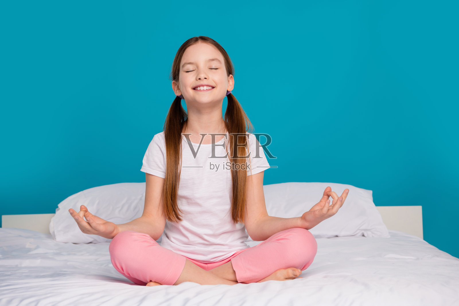 迷人的小女孩的全尺寸照片，闭上眼睛冥想，坐在床上，唤醒卧室，穿着时髦的睡衣，孤立的蓝色背景照片摄影图片