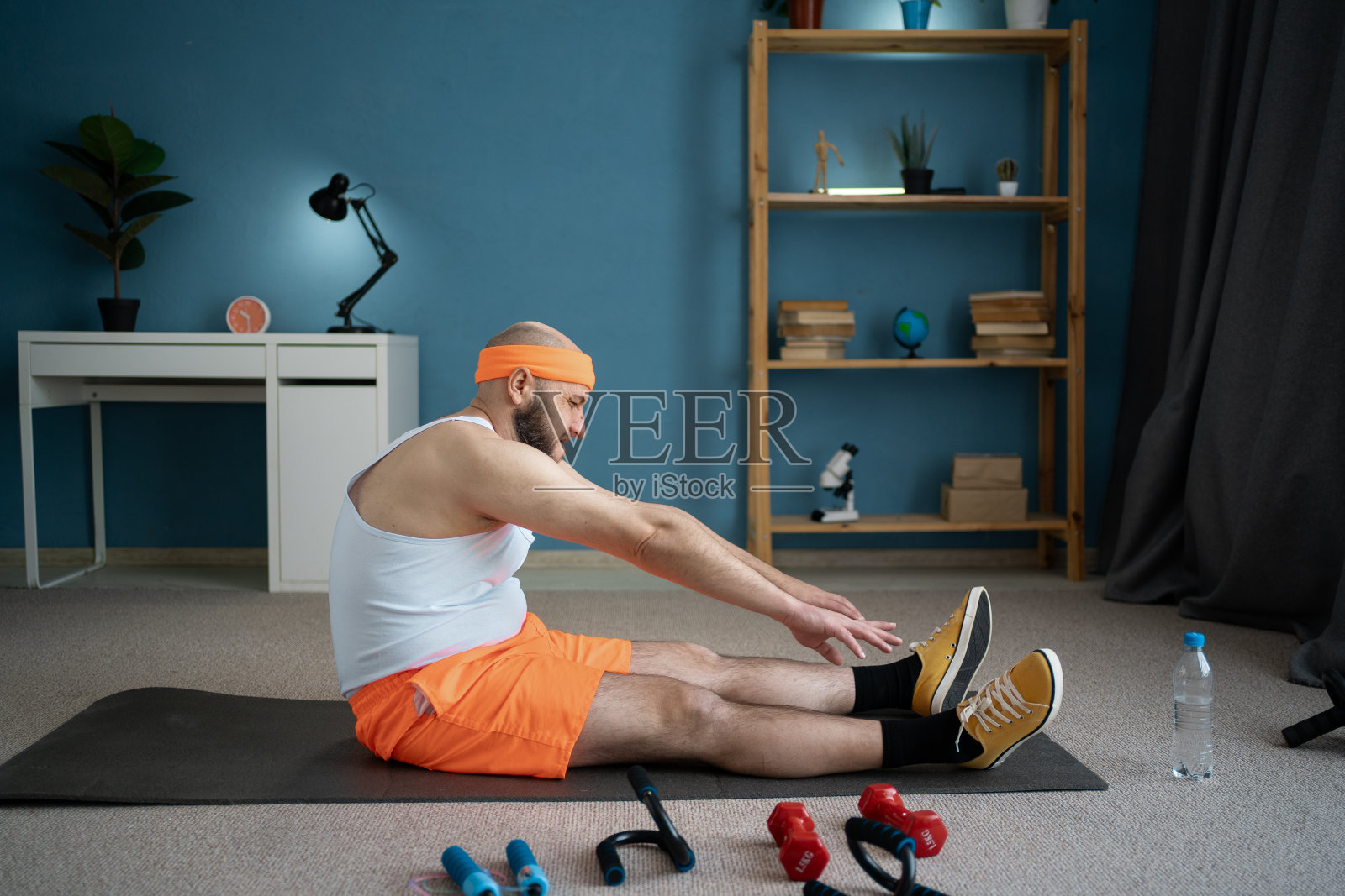 一名男子在家里的瑜伽垫上前屈伸展腿筋。这个房间是为使用运动器材进行家庭锻炼而设置的。家庭健身房照片摄影图片