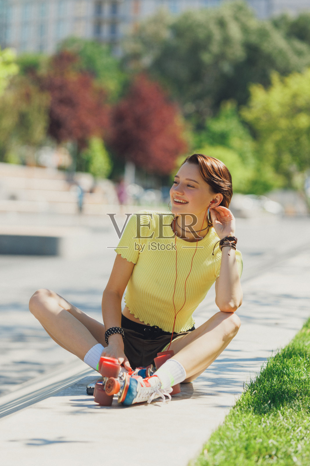 精力充沛，快乐的滑冰女孩享受阳光明媚的公园休息，坐在混凝土坡道上，穿着黄色露膝上衣和棕色短裤。照片摄影图片
