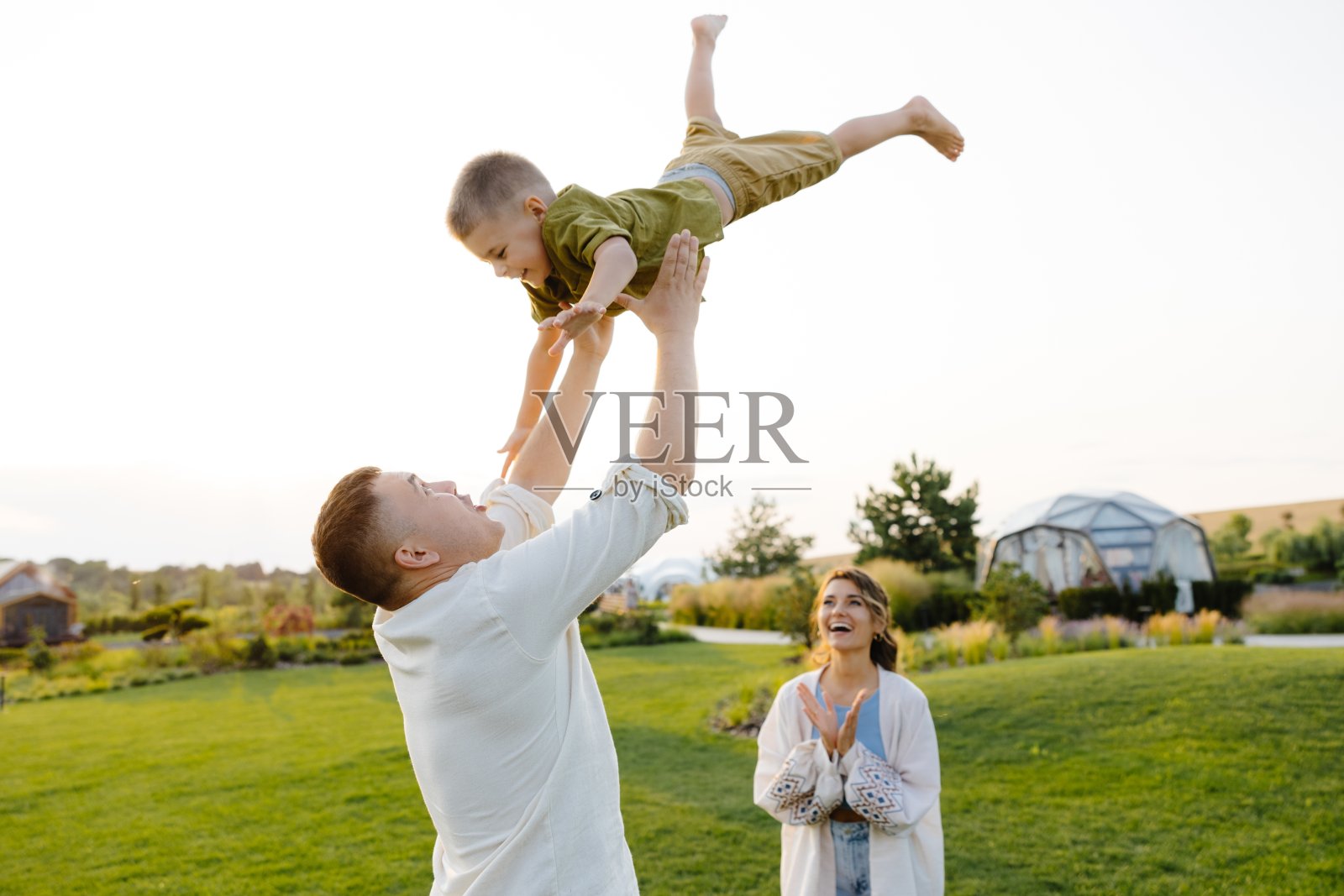 父亲开玩笑地把儿子抛向空中，母亲站在旁边，在美丽的绿色草地上开心地鼓掌微笑照片摄影图片