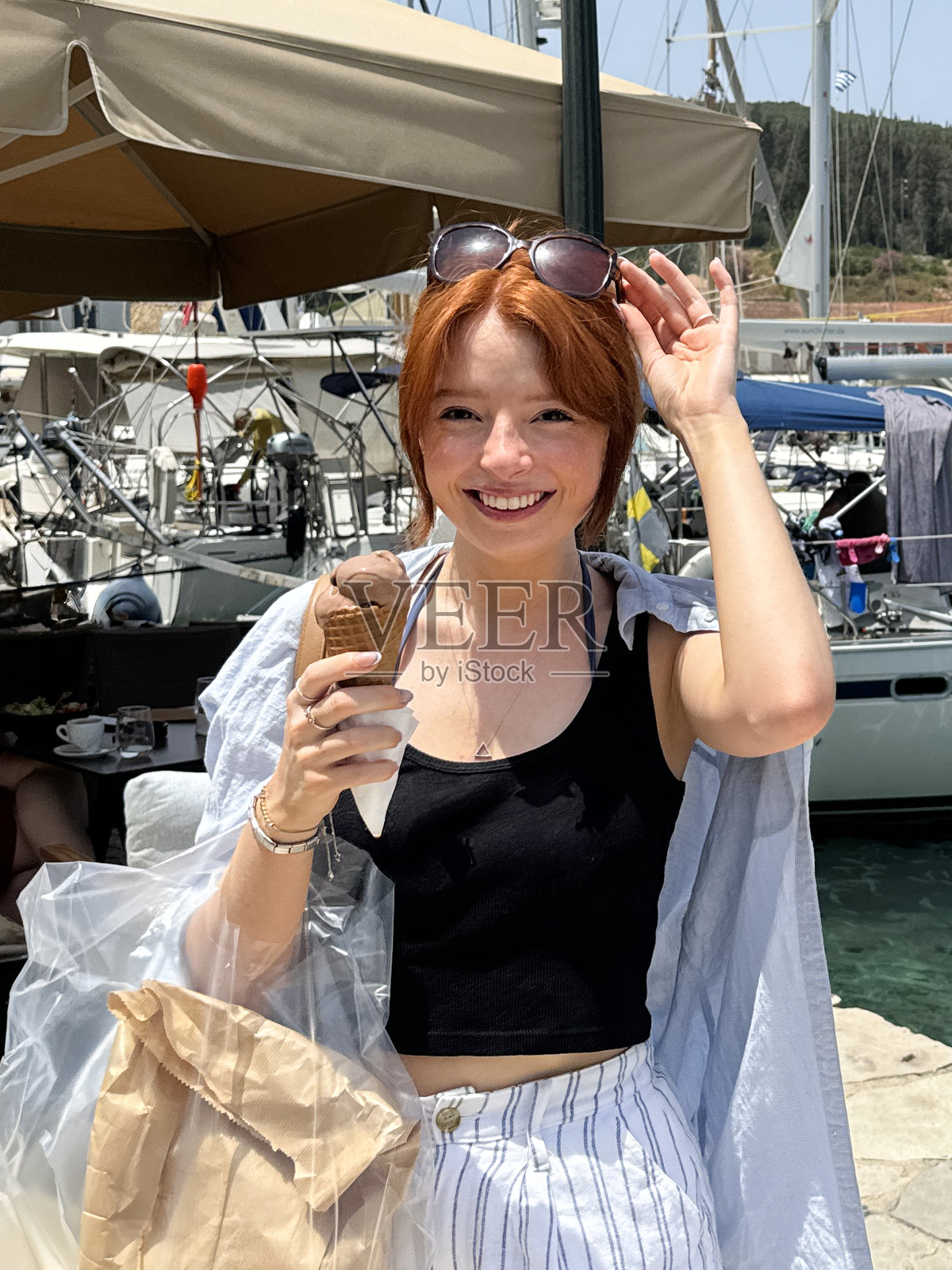 迷人的红发女人头戴墨镜看着相机，拿着含有两勺巧克力冰淇淋的华夫蛋筒，融化的冰淇淋，游艇在码头，港区背景，重点在前景照片摄影图片