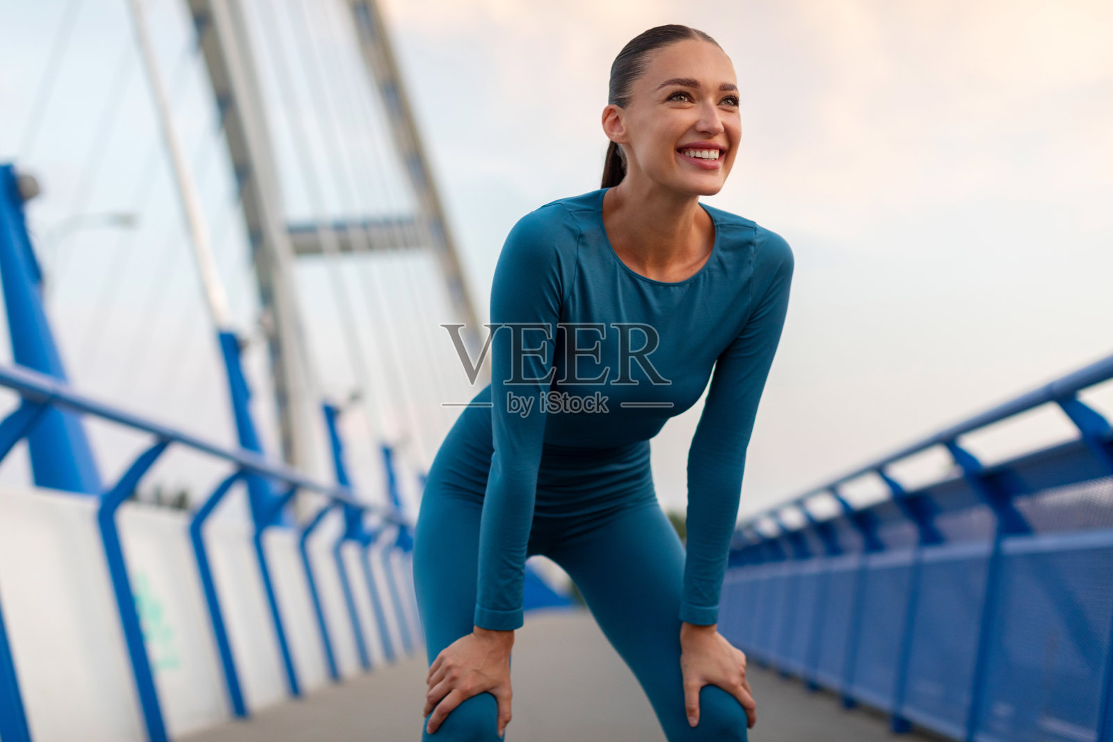 年轻的女运动员在天桥上跑步前靠在膝盖上休息，看着别处，微笑着照片摄影图片