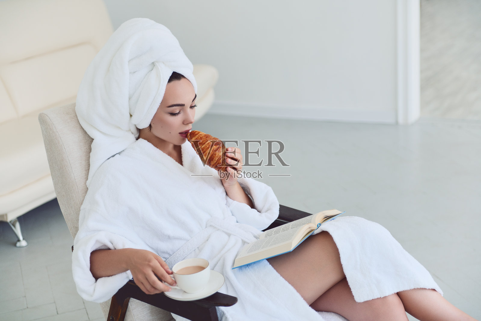 穿着浴袍和毛巾的漂亮女孩坐在椅子上，喝咖啡，吃牛角面包，看书。可爱的年轻女孩早上在家里的画像照片摄影图片