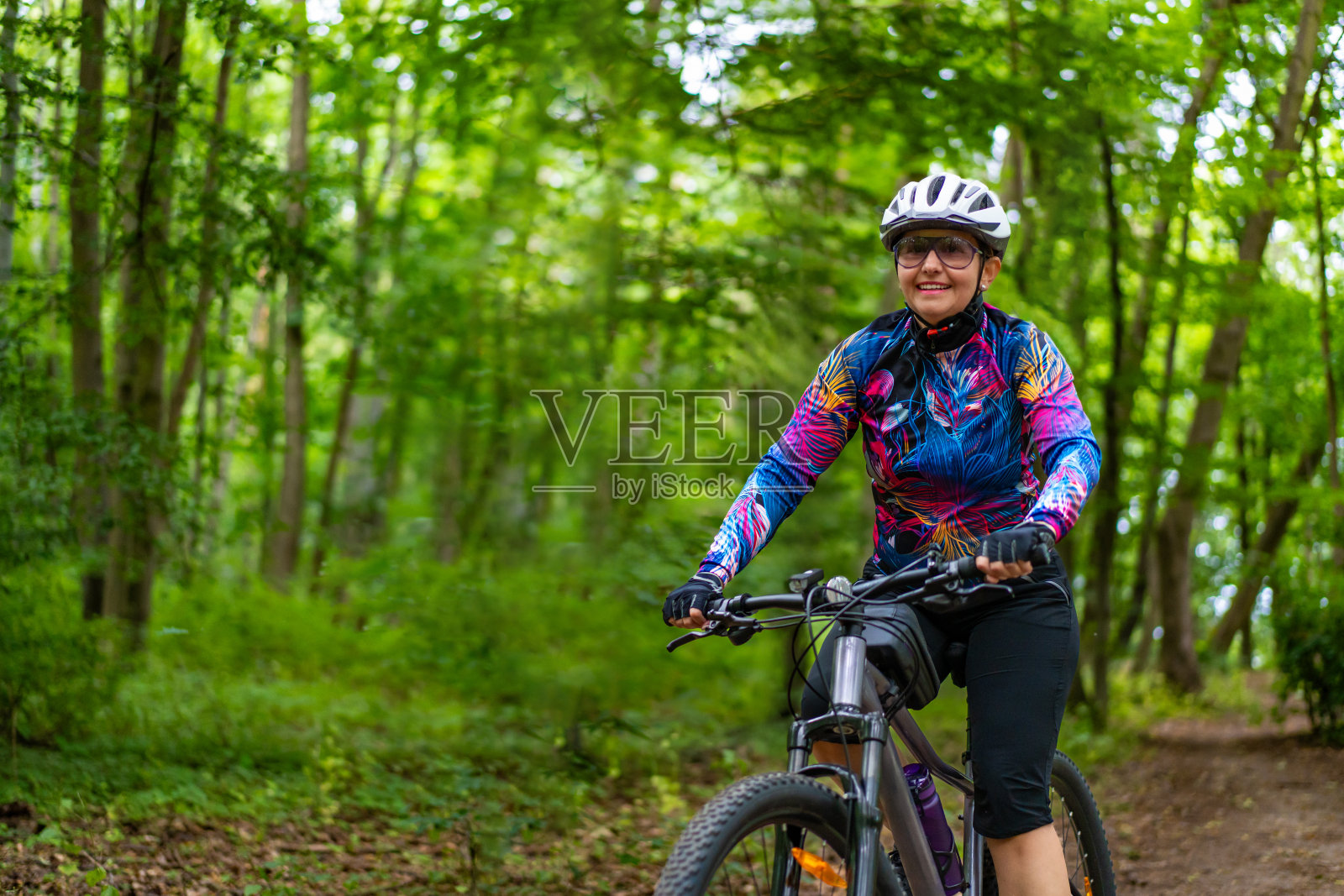 森林骑行训练。戴着白色头盔、穿着彩色衬衫和黑色短裤的年轻女子在森林小路上骑自行车。春天在户外运动。健康的生活方式。前视图照片摄影图片