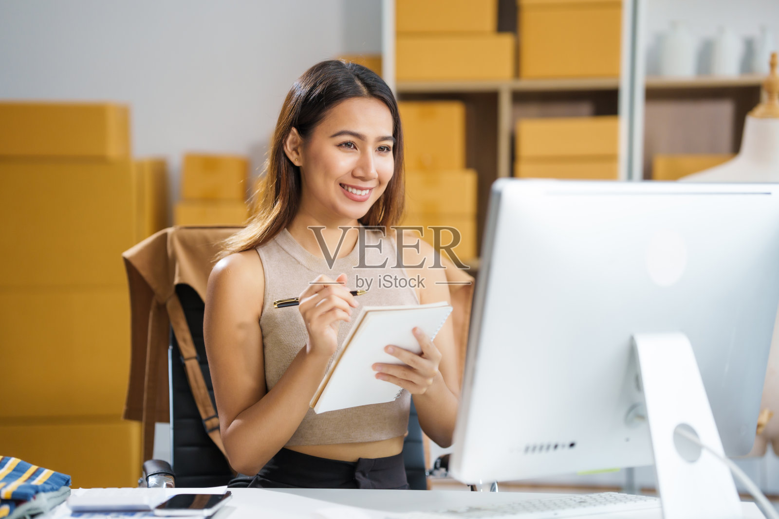 一位年轻的亚洲女商人坐在桌子旁，忙着在网上销售产品。她接受订单，包装物品，并为客户准备货物，准备将它们送到邮局。照片摄影图片