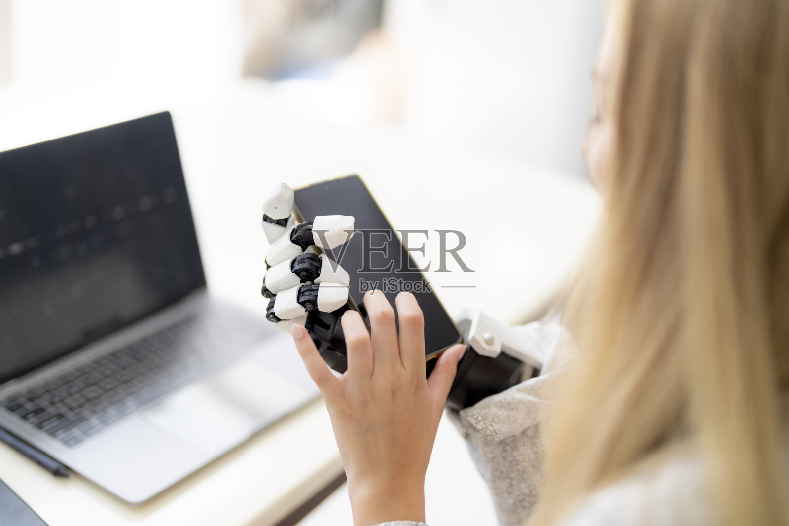 一名年轻女子正在使用附着在她身体上的机械假肢手在桌子上工作。她正在做电脑文件和通讯工作。一位女士试图控制家中的智能手臂。照片摄影图片