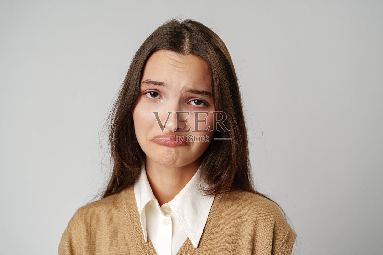 一头棕色长发，身穿白衬衫和棕色毛衣的年轻女子在一堵灰色的墙前看起来很悲伤照片摄影图片