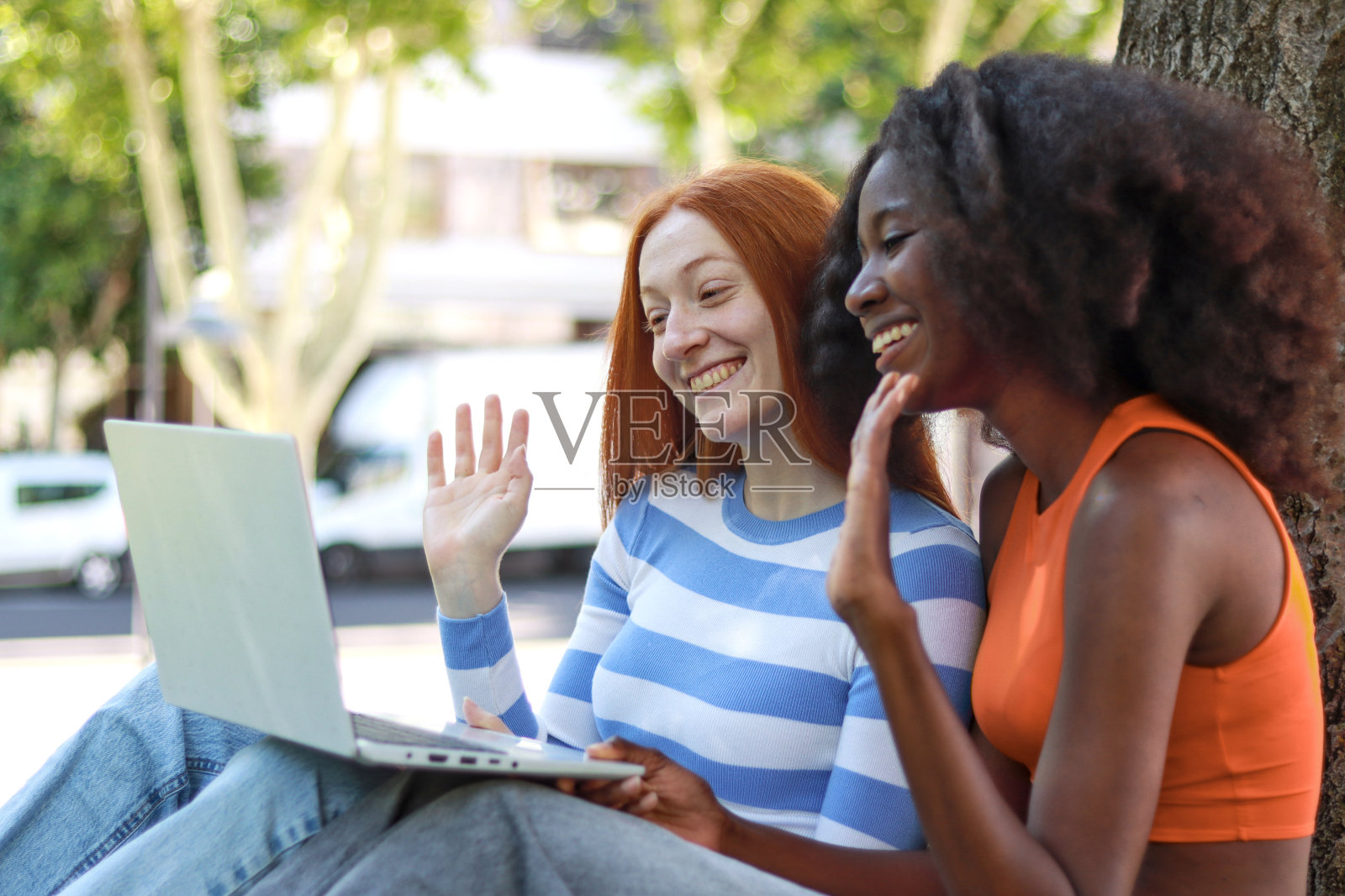 两个年轻女子，一个是非洲人，一个是红发挪威人，她们微笑着挥手，用笔记本电脑进行视频通话照片摄影图片