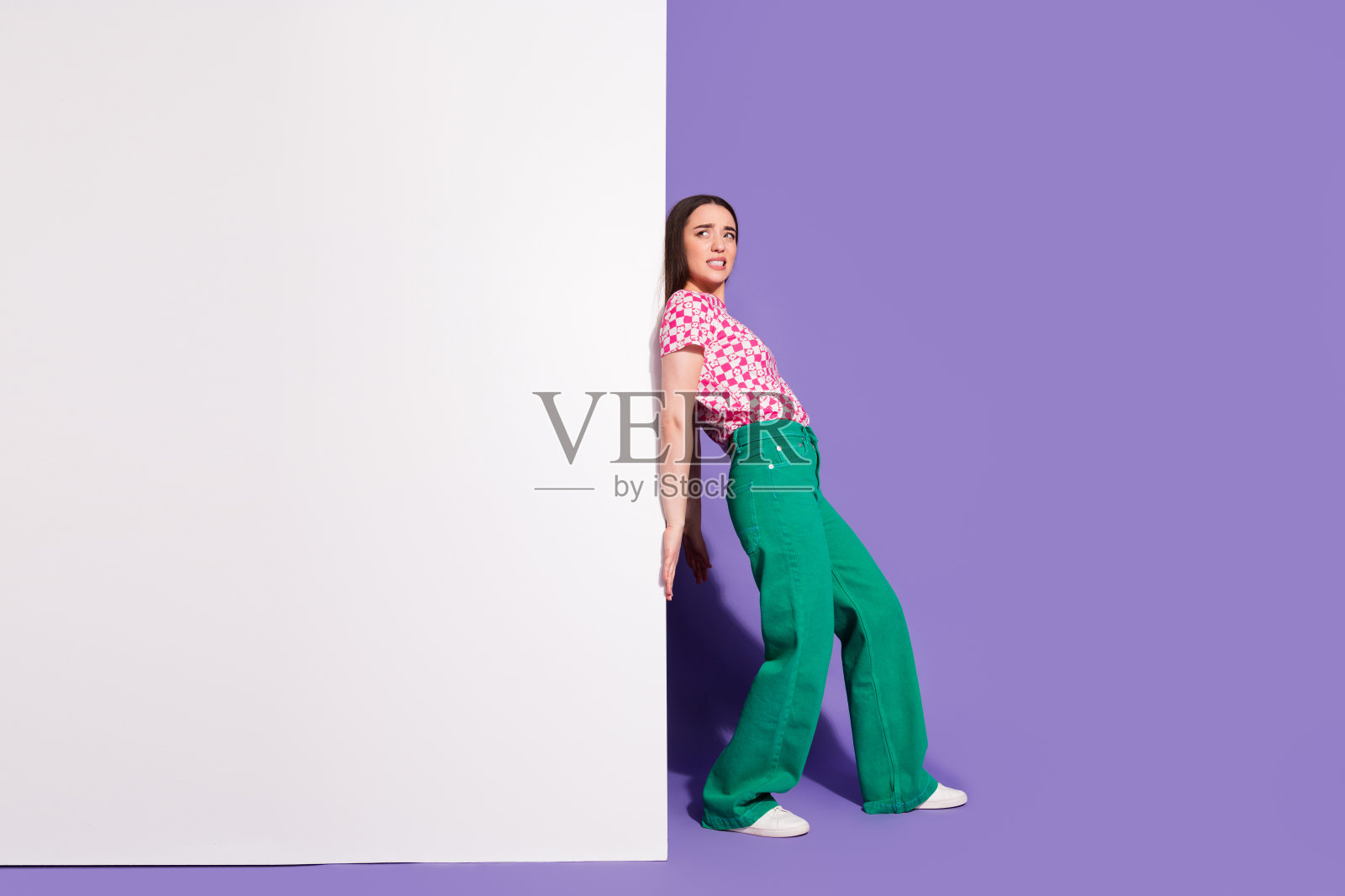 推动受压墙壁、身着时尚粉色服装的可爱年轻女子全身照，紫色背景照片摄影图片