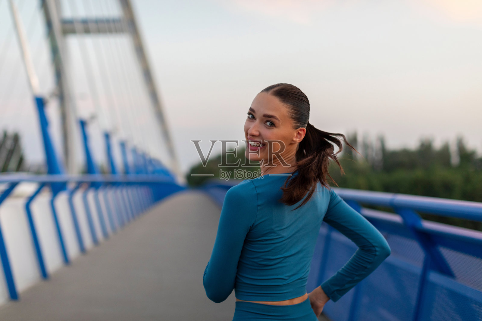 穿着运动服的快乐女人在城市的桥上慢跑，运动健身的女人在户外训练，夕阳下运动，回头微笑照片摄影图片