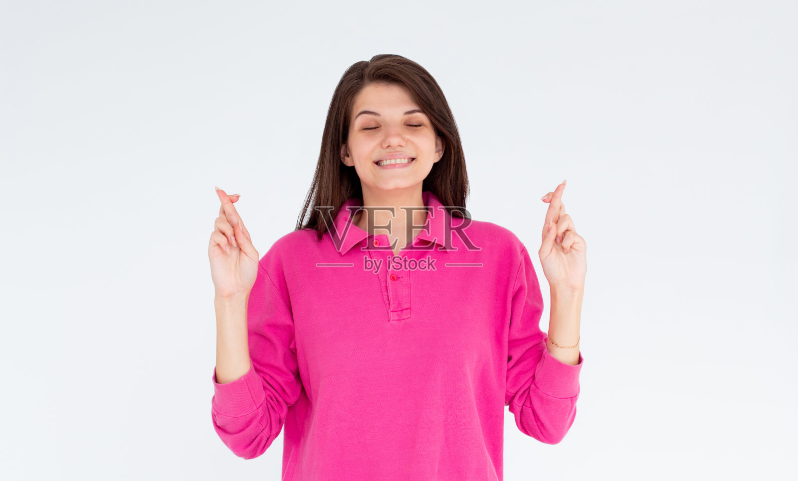 好奇的女人穿着粉红色的长袖，在白色的背景上交叉着手指，闭着眼睛祈祷照片摄影图片