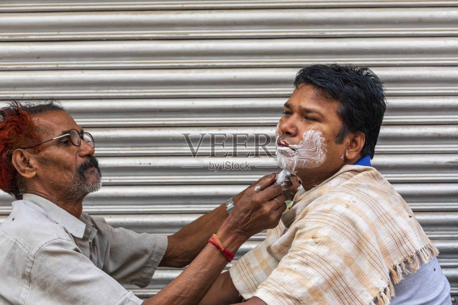 印度，西孟加拉邦，加尔各答，巴拉巴扎尔市场。街头理发师给一个男人刮胡子。照片摄影图片