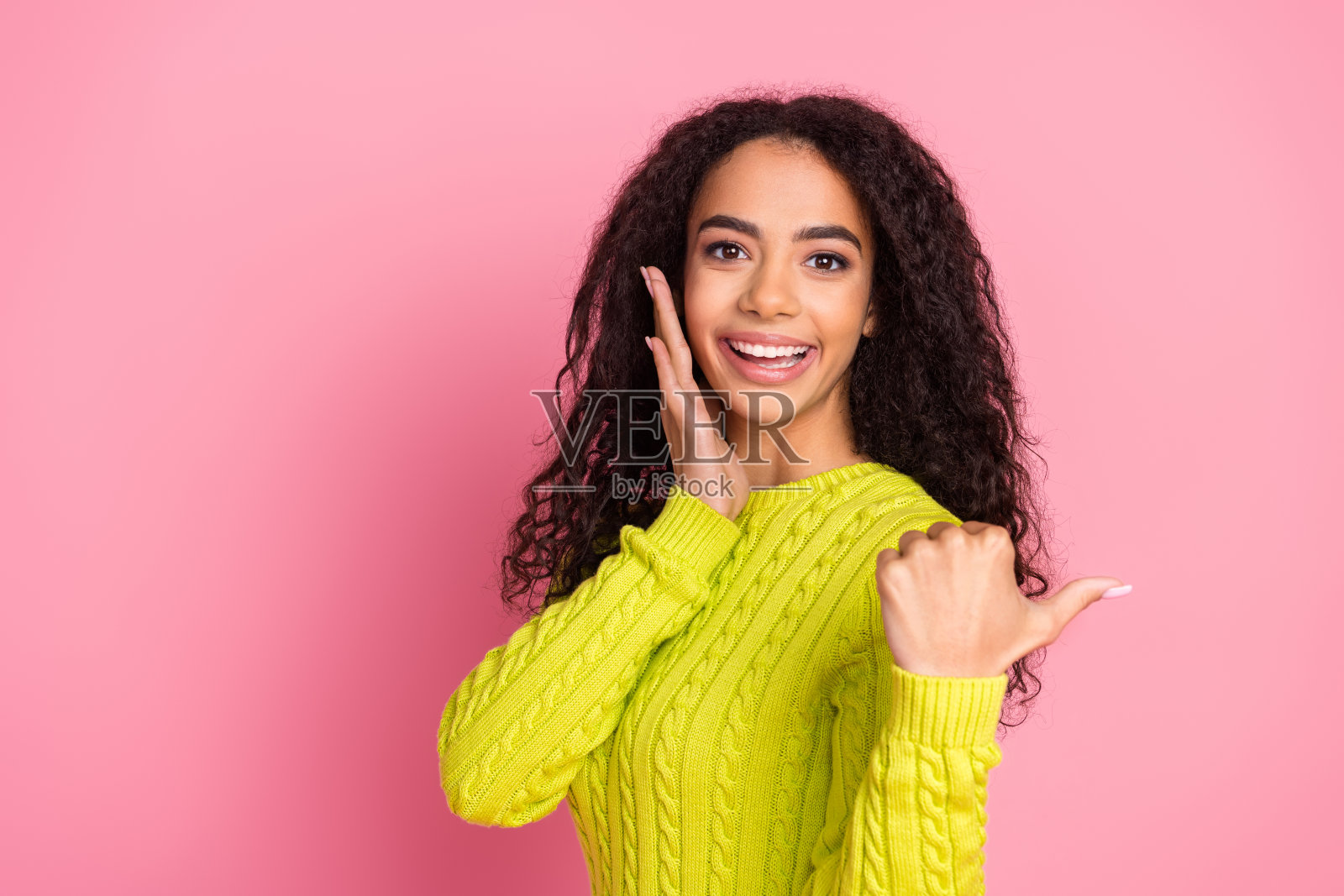 美丽的卷发黑发拉丁女孩的肖像在绿色针织套头衫指向美容院孤立的粉红色背景照片摄影图片